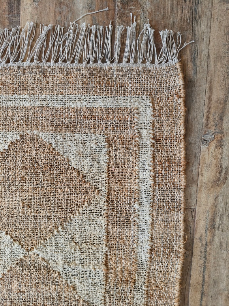 Pu&ograve; includere: Un tappeto in fibra naturale con un motivo geometrico beige e bianco. Il tappeto ha un bordo a frangia.