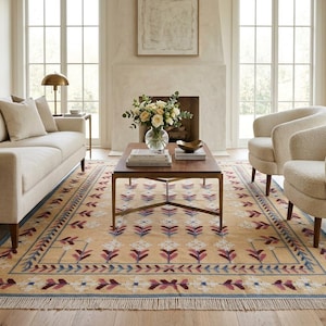 May include: A rectangular area rug featuring a light tan background with a repeating floral pattern in shades of deep red, purple, and blue. The rug has a decorative border and short white fringe along the edges, covering a wooden floor.