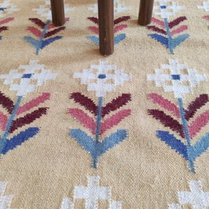 May include: A woven rug with a beige background featuring a repeating pattern of stylized flowers in white, pink, dark red, and blue. Three dark brown wooden legs of a piece of furniture are positioned on the surface of the rug.