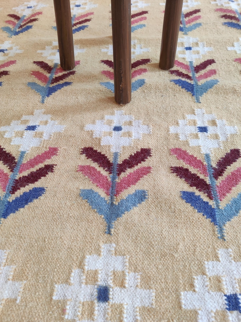 May include: A woven rug with a beige background featuring a repeating pattern of stylized flowers in white, pink, dark red, and blue. Three dark brown wooden legs of a piece of furniture are positioned on the surface of the rug.