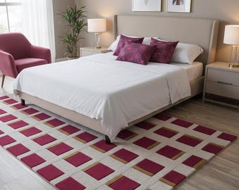 Op de afbeelding: Een slaapkamer met een bed met wit beddengoed en bordeauxrode kussens. Een tapijt met een geometrisch patroon in bordeaux en beige ligt op de vloer. Geometrische kunstwerken sieren de muur boven het bed en een bordeauxrode fauteuil is zichtbaar.