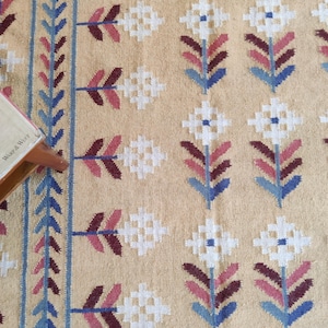 May include: A woven textile featuring a tan background with a repeating pattern of white, maroon, and blue geometric flowers. A vertical border of blue and maroon leaves runs along the left side. The text Warp & Weft is visible on a corner tag.