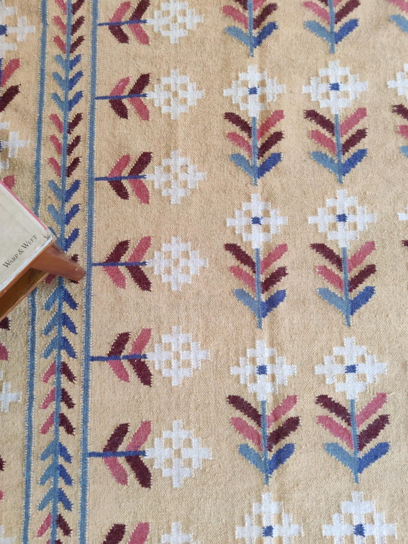 May include: A woven textile featuring a tan background with a repeating pattern of white, maroon, and blue geometric flowers. A vertical border of blue and maroon leaves runs along the left side. The text Warp & Weft is visible on a corner tag.