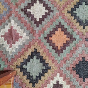 May include: A colorful rug with a geometric pattern in shades of brown, red, green, and white. The rug has a woven texture and a diamond-shaped design.