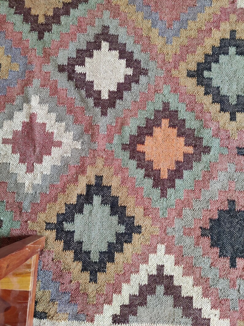May include: A colorful rug with a geometric pattern in shades of brown, red, green, and white. The rug has a woven texture and a diamond-shaped design.