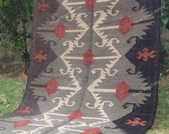 May include: A rectangular rug with a geometric pattern. The rug features a central design in gray, beige, and red, framed by a dark gray border. The rug is made of woven material and has a fringed edge. The text "Floor And Fabrics" is visible.