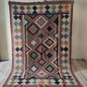 May include: A colorful handwoven rug with a geometric pattern in shades of brown, red, orange, green, and black. The rug has a fringe border.