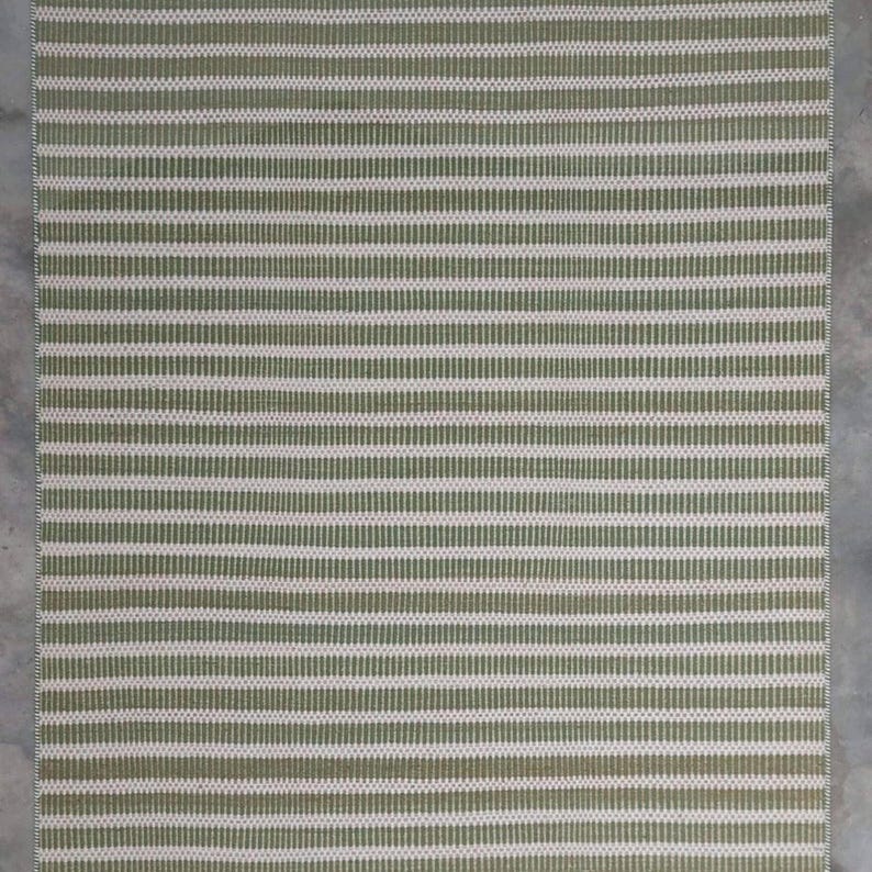May include: A green and white striped woven rug with a subtle texture. The rug has a rectangular shape and is made of natural fibers.