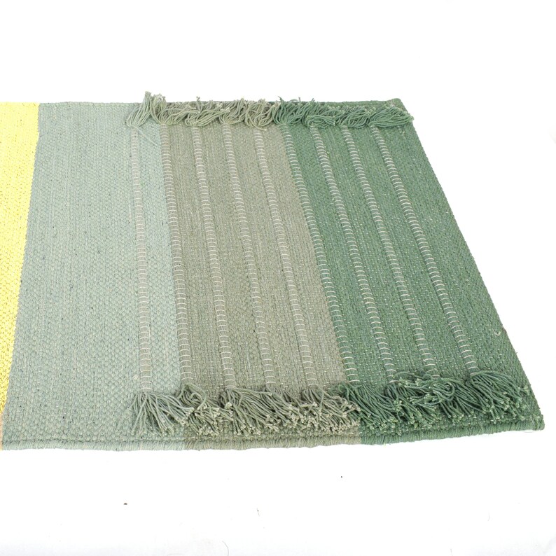 May include: A green and yellow striped rug with a fringe border. The rug is made of a woven material and has a subtle ombre effect.