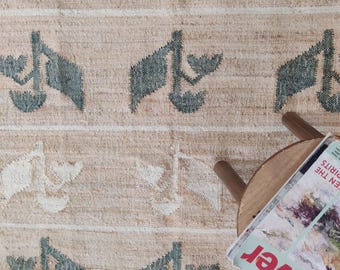 May include: A beige rug with a green and white geometric pattern. The rug is made of a natural fiber and has a slightly textured surface. A wooden stool and a magazine are on top of the rug.