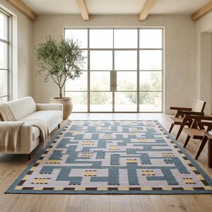 May include: A rectangular area rug featuring a geometric maze-like pattern in shades of slate blue, light gray, and pale yellow, with a dark brown border accent. The rug is positioned on a light wood floor in a room with a sofa and chairs.
