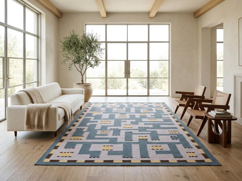 May include: A rectangular area rug featuring a geometric maze-like pattern in shades of slate blue, light gray, and pale yellow, with a dark brown border accent. The rug is positioned on a light wood floor in a room with a sofa and chairs.