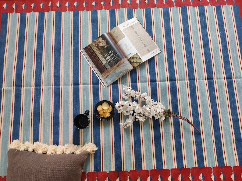 May include: A blue and white striped rug with red accents. The rug has a fringe border and is decorated with a magazine, a bowl of chips, a cup, and a sprig of white flowers.