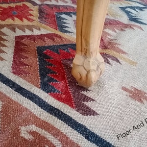 May include: A wooden table leg with a carved claw foot resting on a multi-colored patterned rug. The rug has a geometric design with shades of red, brown, black, and white. The text "Floor And Fabrics" is visible in the lower right corner of the image.