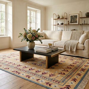 May include: A rectangular area rug featuring a tan base with a repeating pattern of maroon and white floral motifs, framed by a blue border and fringe edges. The rug is positioned under a black coffee table with a vase of flowers and books.