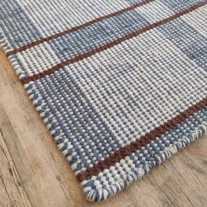 Può includere: Primo piano di un tappeto tessuto con un motivo a quadri blu e bianco. Il tappeto presenta strisce marroni e un bordo sfrangiato. Il tappeto è realizzato in un materiale strutturato ed è posizionato su una superficie di legno.