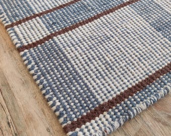 Puede incluir: Primer plano de una alfombra tejida con un patrón de cuadros azules y blancos. La alfombra presenta rayas marrones y un borde con flecos. La alfombra está hecha de un material texturizado y está colocada sobre una superficie de madera.