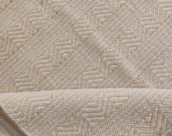 May include: A close-up of a beige woven rug with a chevron pattern. The rug is folded in half, showing the texture of the weave.