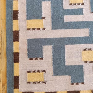 May include: A woven rug featuring a geometric maze pattern in slate blue, light gray, and yellow. The border consists of a repeating checkerboard design in alternating dark brown and yellow squares, framing the central angular, interlocking shapes.