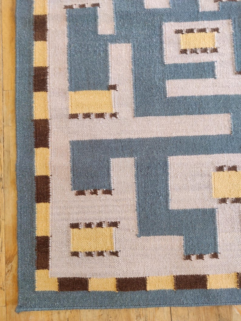 May include: A woven rug featuring a geometric maze pattern in slate blue, light gray, and yellow. The border consists of a repeating checkerboard design in alternating dark brown and yellow squares, framing the central angular, interlocking shapes.