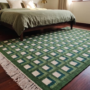 May include: A rectangular area rug featuring a repeating geometric pattern of nested squares in shades of forest green, sage green, and cream. The rug has a textured pile and is finished with long, cream-coloured fringe along the short edges.