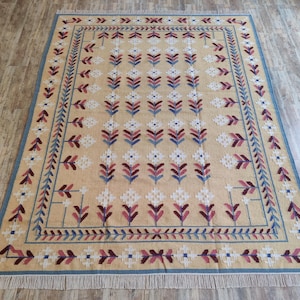 May include: A rectangular area rug featuring a tan background with a repeating pattern of white, blue, and maroon floral motifs. The rug is framed by a blue border with a geometric leaf design and finished with white fringe on both short ends.