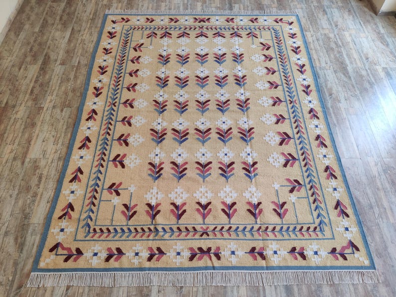 May include: A rectangular area rug featuring a tan background with a repeating pattern of white, blue, and maroon floral motifs. The rug is framed by a blue border with a geometric leaf design and finished with white fringe on both short ends.