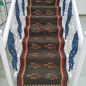 以下が含まれることがあります： 赤と金のアクセントが付いた、濃い茶色と黒の模様の敷物。敷物はフリンジの縁取りがあり、白と青の手すりの付いた階段に敷かれています。