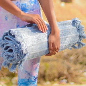 May include: A person holding a rolled up light blue yoga mat with fringe. The mat is made of a soft, textured fabric.
