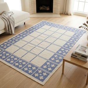 May include: A rectangular area rug featuring a cream-colored center with a grid pattern and a wide border composed of blue squares, each containing a white cross motif. The rug has a flat-woven texture and a repeating geometric design throughout.