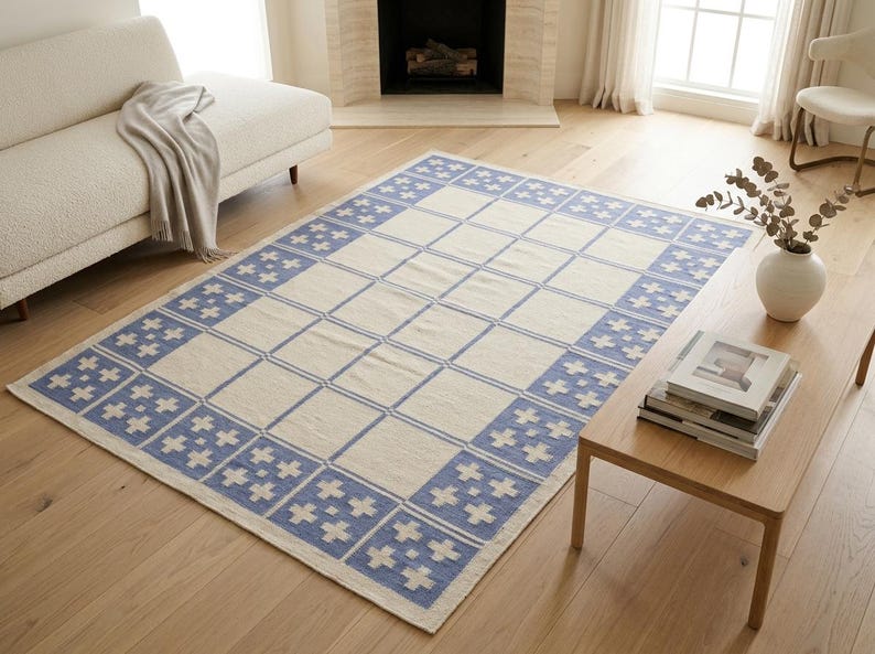 May include: A rectangular area rug featuring a cream-colored center with a grid pattern and a wide border composed of blue squares, each containing a white cross motif. The rug has a flat-woven texture and a repeating geometric design throughout.