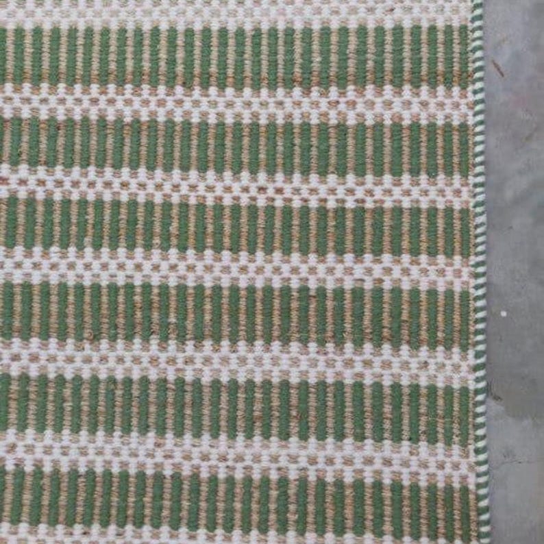 May include: A green and white striped woven rug with a subtle texture. The rug has a simple design and is made of natural fibers.