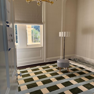 May include: An interior room with a patterned rug in shades of green, blue, and beige. A small gray stool and a tall lamp stand near the window. A gold chandelier hangs from the ceiling, and the window has a patterned shade.