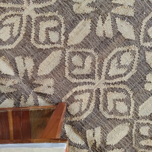May include: A close-up of a brown woven rug with a white geometric pattern. The rug has a textured surface and is made of natural fibers. A wooden table leg is visible in the lower left corner of the image.