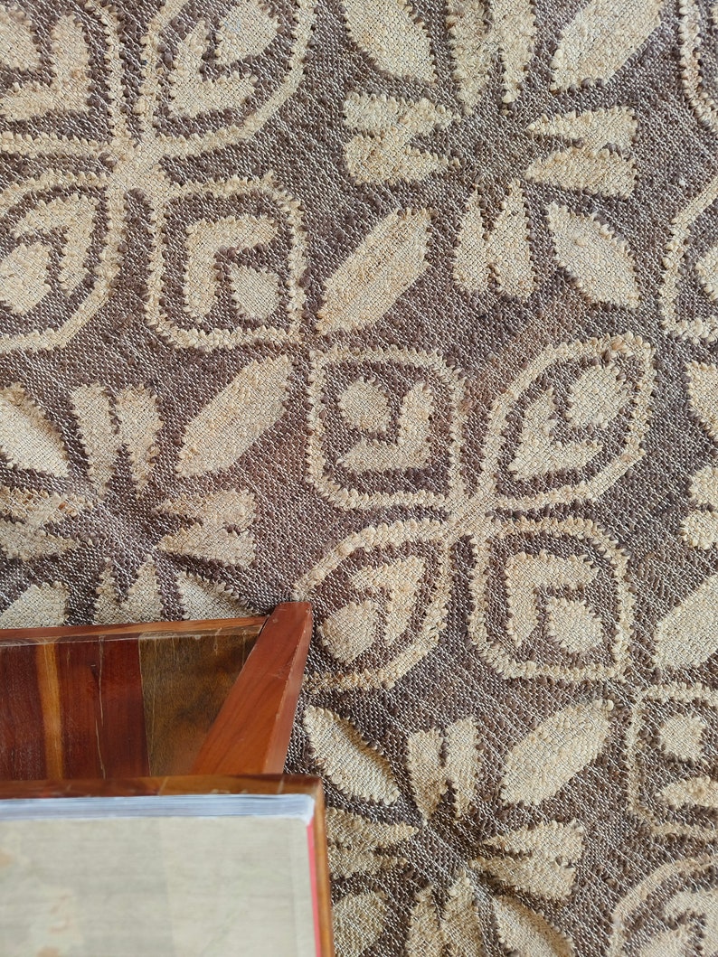 May include: A close-up of a brown woven rug with a white geometric pattern. The rug has a textured surface and is made of natural fibers. A wooden table leg is visible in the lower left corner of the image.