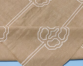 Puede incluir: Tela beige con un patrón floral geométrico blanco repetido y rayas blancas diagonales. La tela está doblada, mostrando una esquina. El fondo es azul claro.