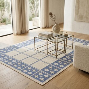 May include: A rectangular area rug featuring a cream-colored center and a wide border decorated with a repeating blue and cream cross pattern. The rug is positioned on a light wood floor, partially underneath a set of three nested glass tables.