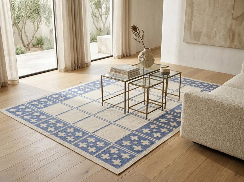 May include: A rectangular area rug featuring a cream-colored center and a wide border decorated with a repeating blue and cream cross pattern. The rug is positioned on a light wood floor, partially underneath a set of three nested glass tables.