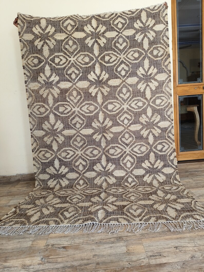 May include: A brown and cream patterned rug with a fringe border. The rug has a geometric design with a repeating pattern of stylized flowers.
