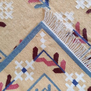 May include: A beige woven rug featuring a repeating pattern of white cross shapes, dark red and pink leaf-like motifs, and blue geometric lines. The rug has a folded corner revealing a blue border and white fringe along the edge of the fabric.