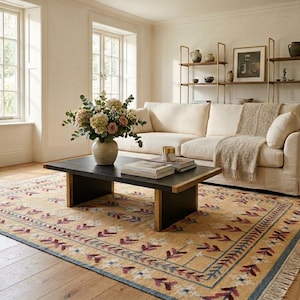 May include: A rectangular area rug featuring a tan base with a repeating pattern of maroon and white floral motifs, framed by a blue border and fringe edges. The rug is positioned under a black coffee table with a vase of flowers and books.