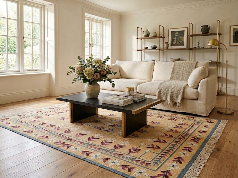 May include: A rectangular area rug featuring a tan base with a repeating pattern of maroon and white floral motifs, framed by a blue border and fringe edges. The rug is positioned under a black coffee table with a vase of flowers and books.