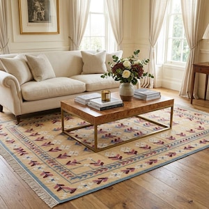 May include: A rectangular area rug featuring a light tan background with a repeating pattern of stylized floral motifs in shades of deep red, white, and blue. The rug has a decorative border and short fringe along the edges, laid on a wooden floor.