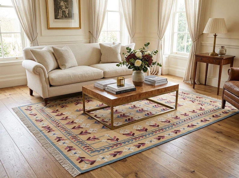 May include: A rectangular area rug featuring a light tan background with a repeating pattern of stylized floral motifs in shades of deep red, white, and blue. The rug has a decorative border and short fringe along the edges, laid on a wooden floor.