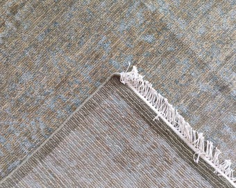 May include: A textured rug with a corner folded back, showing the underside. The rug has a pattern of light blue and tan colours. The edge has a white fringe.