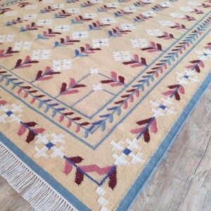 May include: A rectangular area rug with a beige background featuring a repeating pattern of white, blue, and maroon floral motifs. The rug has a blue border with a leaf design and white fringe along the short edges, creating a textured surface.