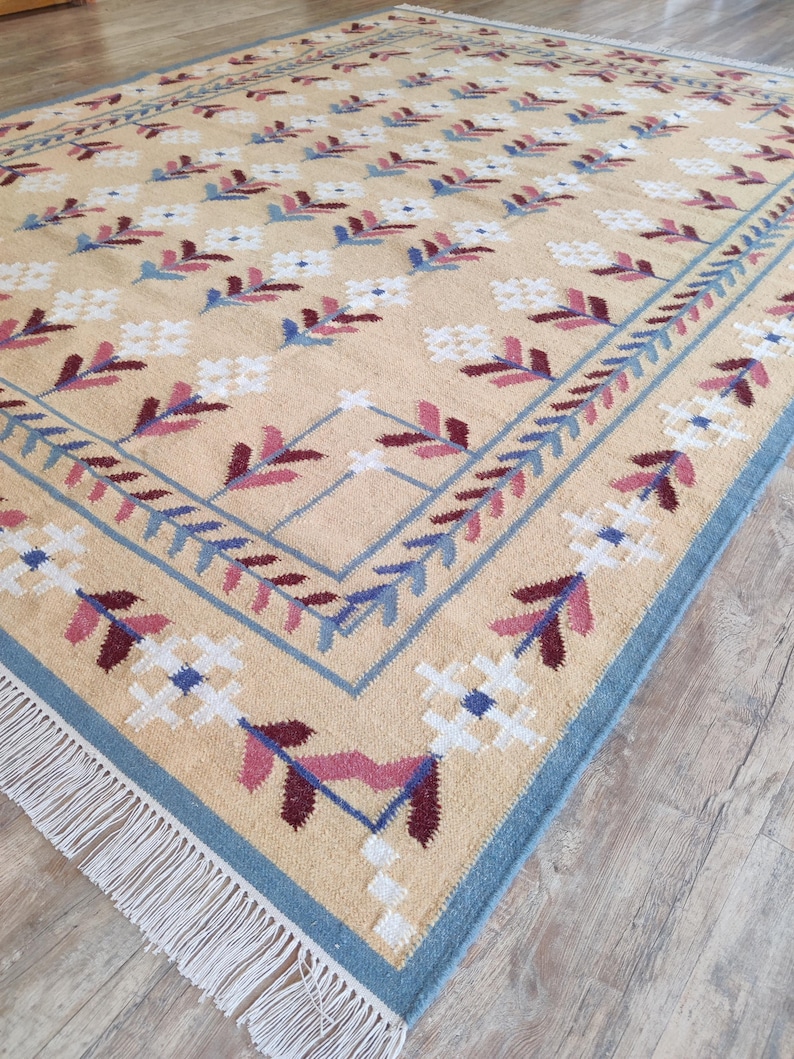 May include: A rectangular area rug with a beige background featuring a repeating pattern of white, blue, and maroon floral motifs. The rug has a blue border with a leaf design and white fringe along the short edges, creating a textured surface.