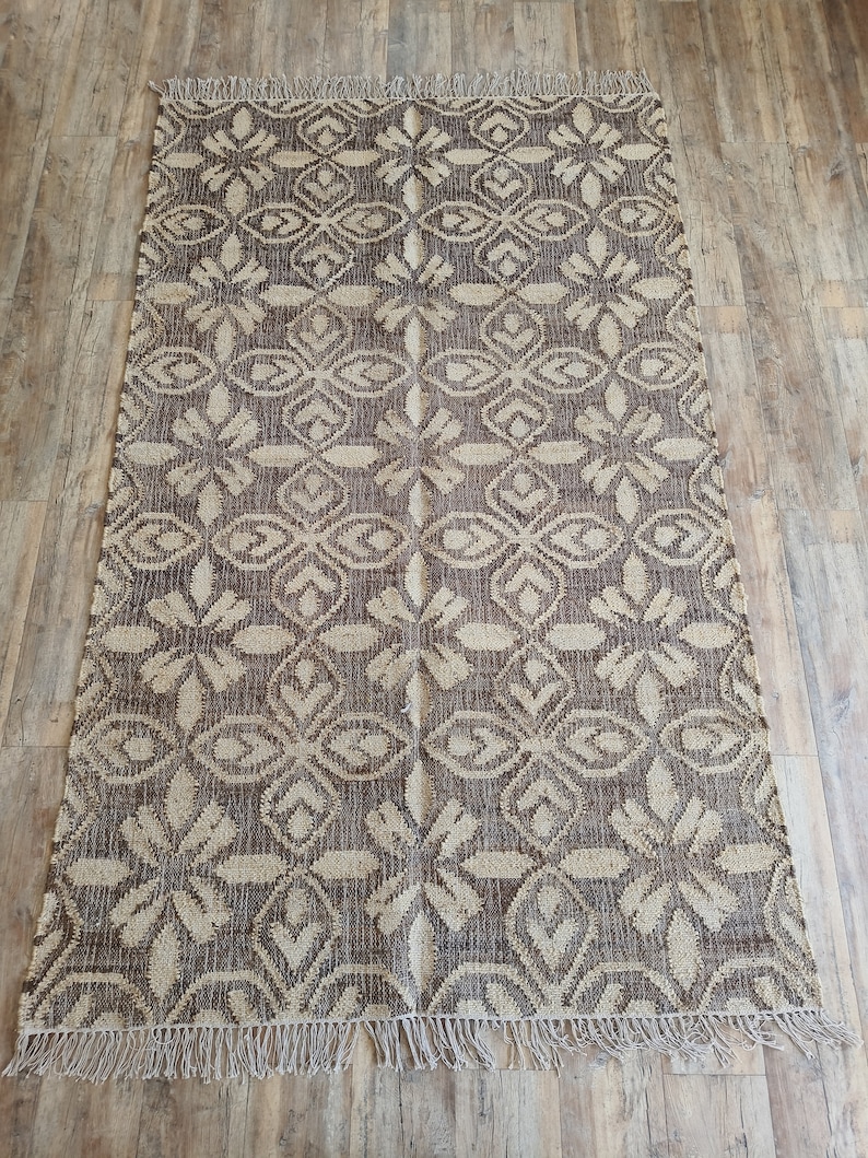 May include: A brown and beige patterned rug with a fringe border. The rug has a geometric design with a repeating pattern of diamonds and flowers.