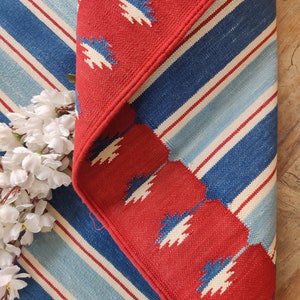 May include: Red and blue striped rug with a red border featuring a white geometric pattern. The rug has a fringe border.