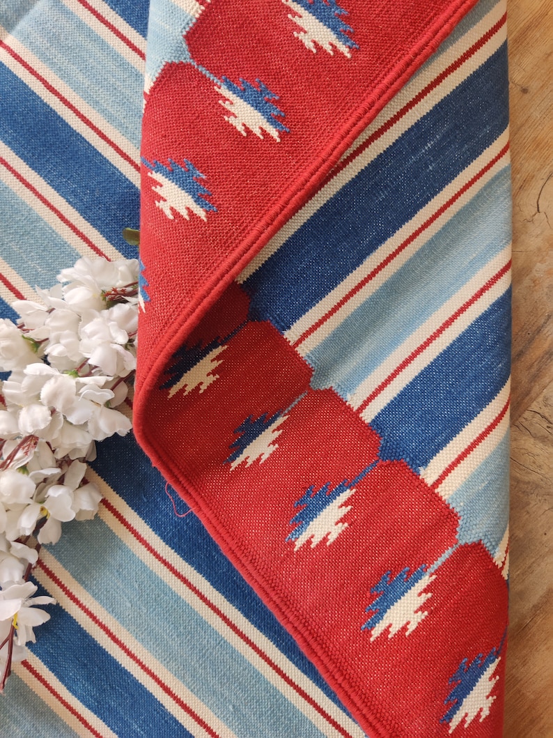 May include: Red and blue striped rug with a red border featuring a white geometric pattern. The rug has a fringe border.